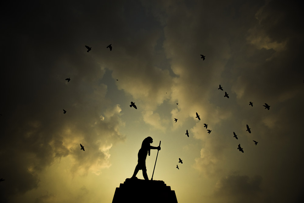 birds fly over a statue of Mahatma Gandhi, silhouetted against the evening sky on the eve of his birth anniversary in Amritsar