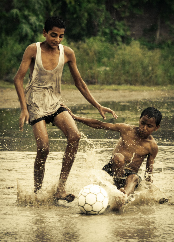 young boys play soccer in rain water puddle.